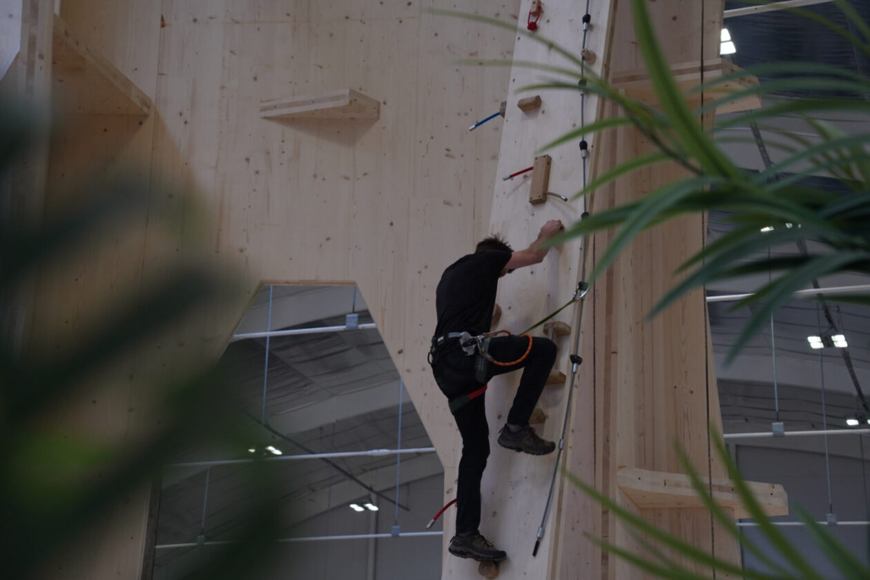 Climber scaling a tall wooden indoor climbing wall with holds and rope access gear visible at the waist.