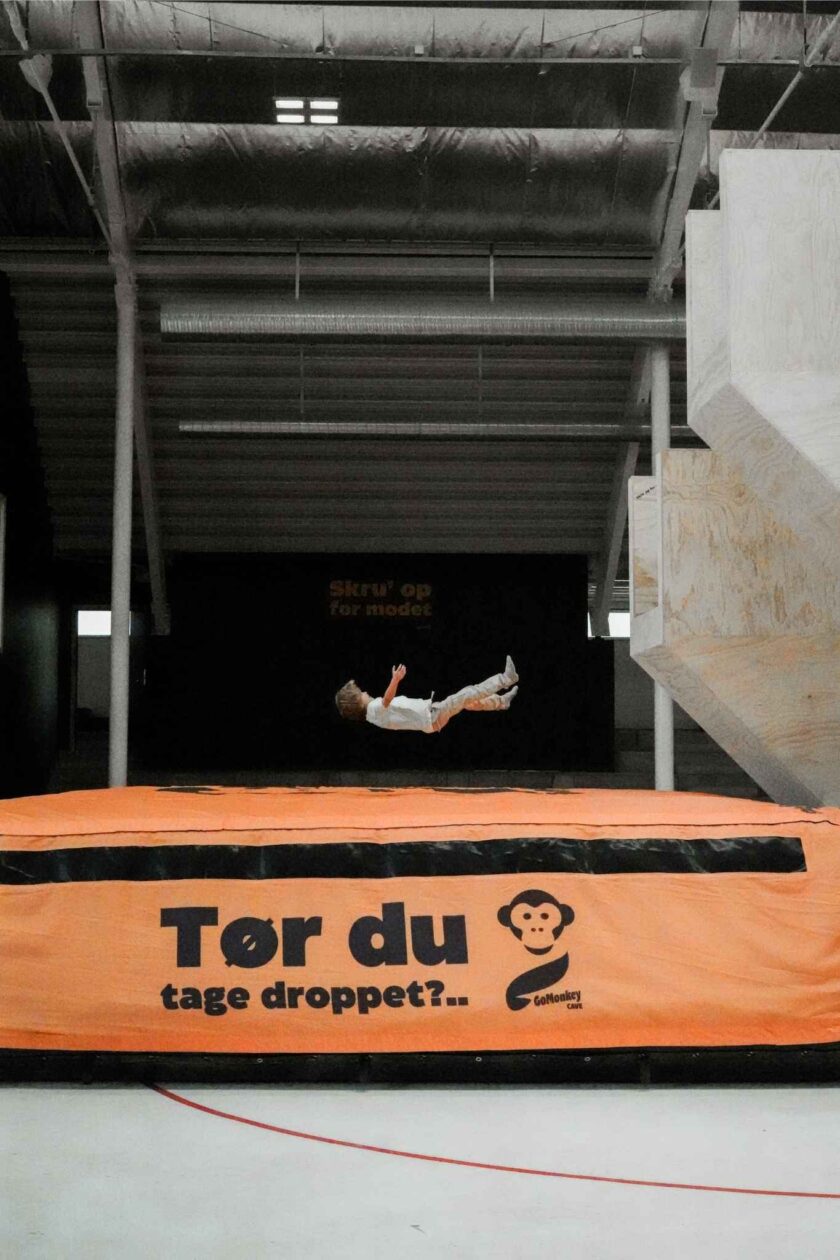A child mid-air performing a jump on an orange-trampoline arena indoors, with a black backdrop and exposed ceiling.